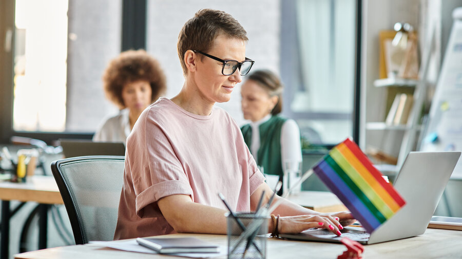 Eine Frau arbeitet im Büro am Computer. Neben ihr steht eine Regenbogenfahne auf dem Schreibtisch. 