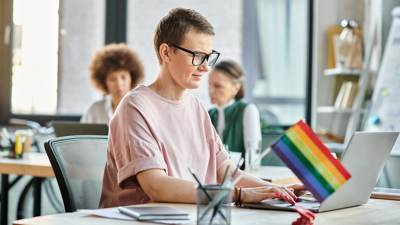 Eine Frau arbeitet im Büro am Computer. Neben ihr steht eine Regenbogenfahne auf dem Schreibtisch. 