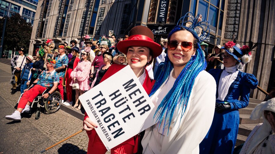 Zu sehen sind zwei Frauen, die freundlich in die Kamera schauen. Sie halten ein Schild, auf dem steht: Mehr Hüte tragen. Es handelt sich um ein Foto des World Head Walk 2025.