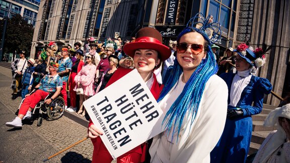 Zu sehen sind zwei Frauen, die freundlich in die Kamera schauen. Sie halten ein Schild, auf dem steht: Mehr Hüte tragen. Es handelt sich um ein Foto des World Head Walk 2025.
