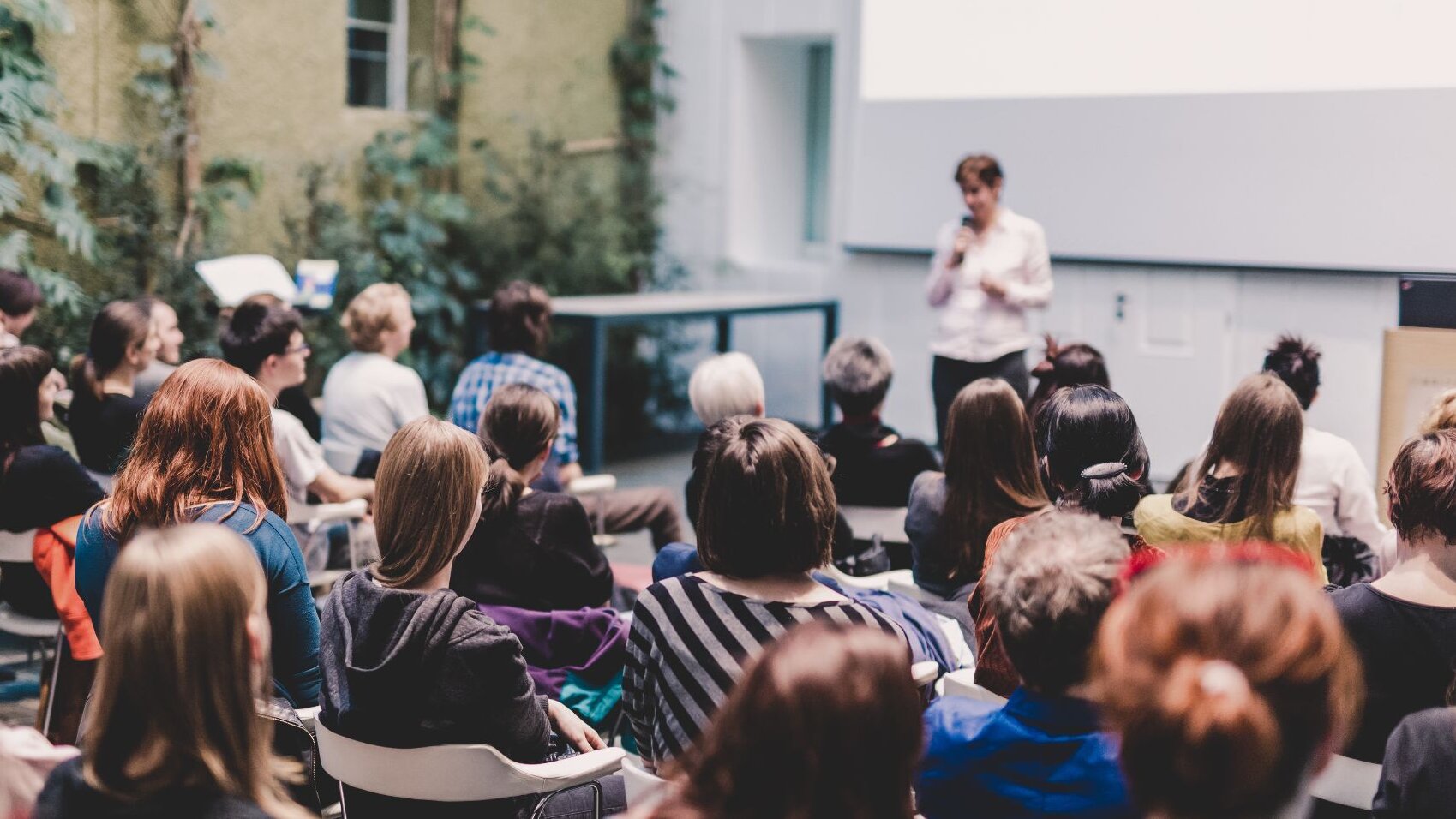 Zu sehen ist sitzendes Publikum. Dieses besteht aus Frauen, welche von hinten zu sehen sind. Im Hintergrund eine Bühne, mit einer unscharf zu erkennenden Frau, die zum Publikum spricht.