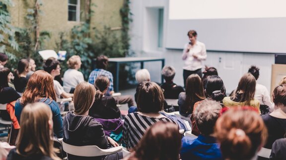 Zu sehen ist sitzendes Publikum. Dieses besteht aus Frauen, welche von hinten zu sehen sind. Im Hintergrund eine Bühne, mit einer unscharf zu erkennenden Frau, die zum Publikum spricht.