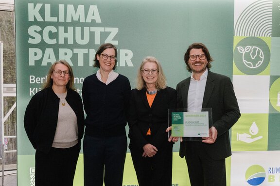 Zu sehen sind vier Personen, ein Mann und drei Frauen, die vor einer Fotowand stehen und freundlich in die Kamera schauen.