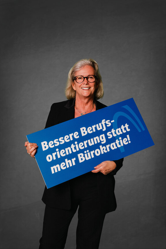 Eine Person steht vor einem neutralen grauen Hintergrund und hält ein großes blaues Schild mit der weißen Aufschrift: "Bessere Berufsorientierung statt mehr Bürokratie!""