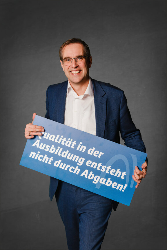 Eine Person steht vor einem neutralen grauen Hintergrund und hält ein großes blaues Schild mit der weißen Aufschrift: "Qualität in der Ausbildung entsteht nicht durch Abgaben!"