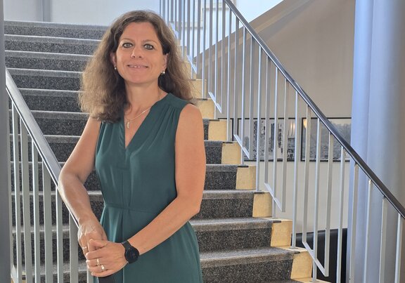 Zu sehen ist eine Frau mit dunkelbraunen Haaren in einem grünen Kleid auf einer Treppe