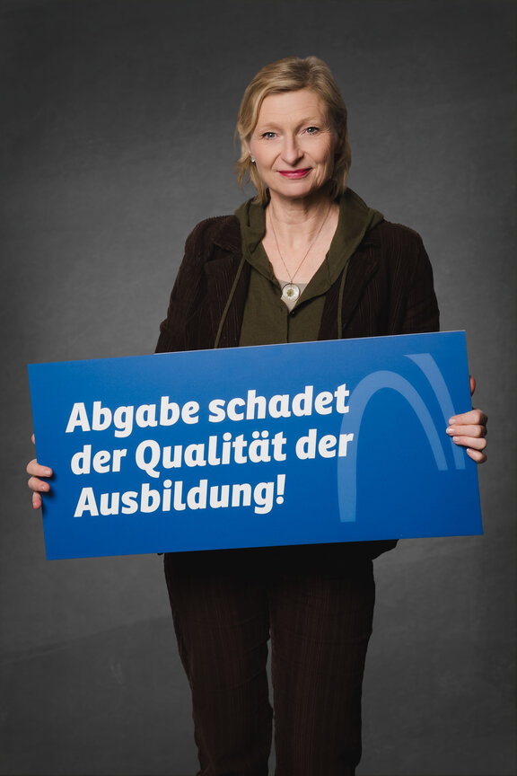Eine Person steht vor einem neutralen grauen Hintergrund und hält ein großes blaues Schild mit der weißen Aufschrift: "Abgabe schadet der Qualität der Ausbildung!"