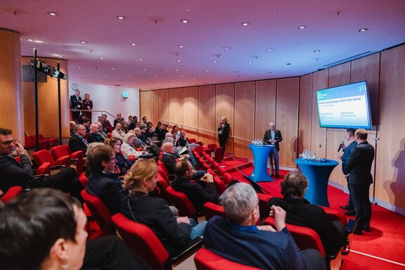 Blick von hinten über das Publikum auf die Bühne einer Podiumsdiskussion.