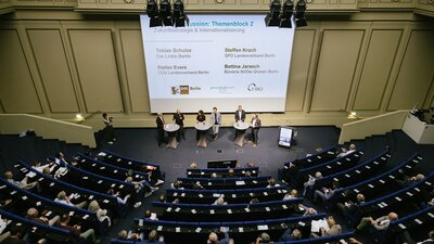 Ein Auditorium mit Gästen des wahlpolitischen Dialogs und die Diskutanten vor einer Leinwand