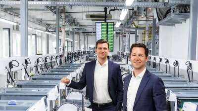 Zu sehen sind zwei Männer im Anzug, die in einer Produktions-/Industriehalle stehen und freundlich in die Kamera schauen.