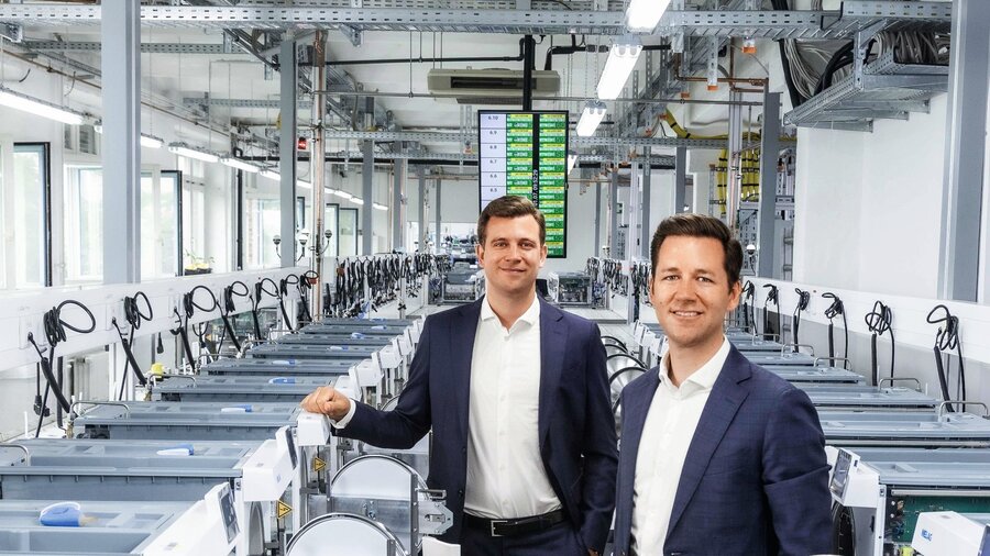 Zu sehen sind zwei Männer im Anzug, die in einer Produktions-/Industriehalle stehen und freundlich in die Kamera schauen.