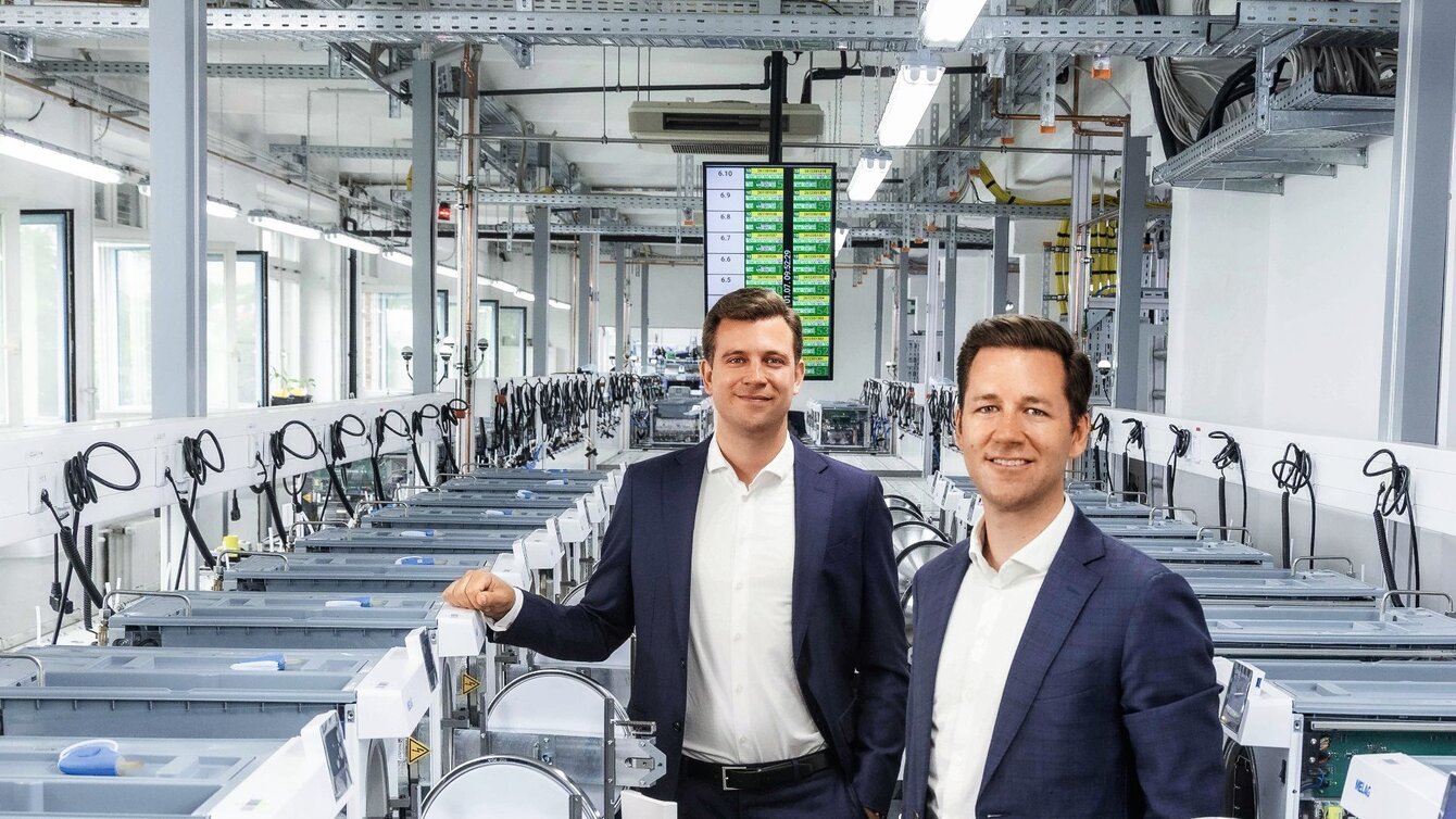 Zu sehen sind zwei Männer im Anzug, die in einer Produktions-/Industriehalle stehen und freundlich in die Kamera schauen.