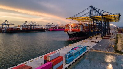 Zu sehen ist ein großes Güterschiff und zahlreiche Container am Wasser.
