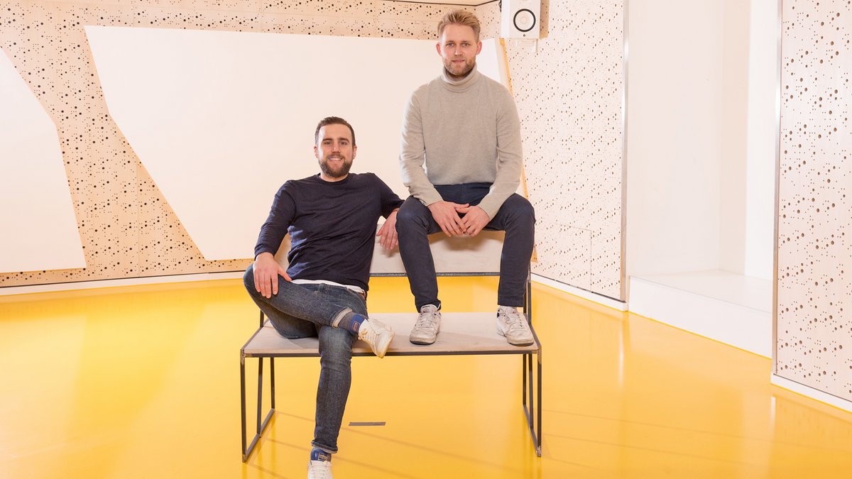 Zwei Männer in lässiger Kleidung sitzen auf einem Tisch in einem Raum mit gelbem Boden und weiß-beiger Wandgestaltung.