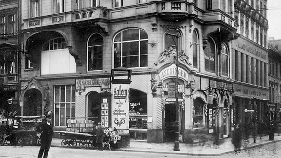 Historische Fassade des Restaurants Aschinger in einer belebten Straßenecke mit Passanten und Pferdekutschen