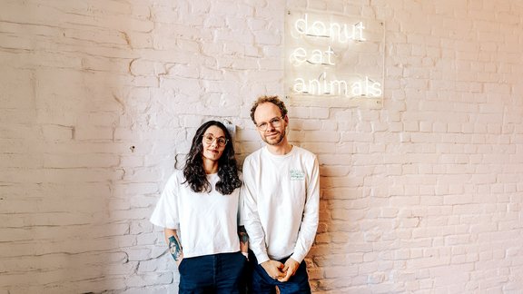 Zwei Personen stehen vor einer weißen Backsteinwand mit leuchtendem Schild 'donut eat animals'