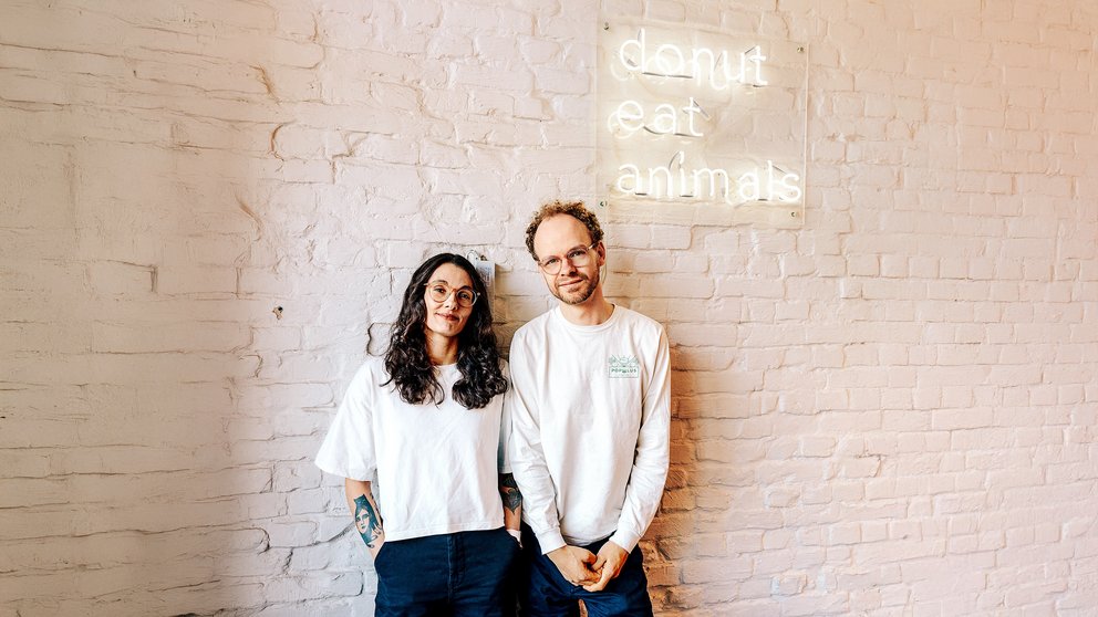 Zwei Personen stehen vor einer weißen Backsteinwand mit leuchtendem Schild 'donut eat animals'
