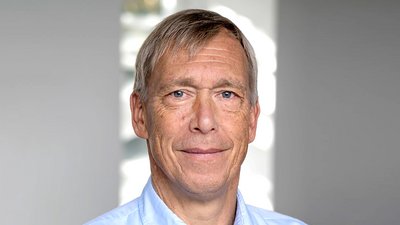 Mann mit grauen Haaren trägt ein hellblaues Hemd und steht vor einem hellen Hintergrund mit Fensterlicht.