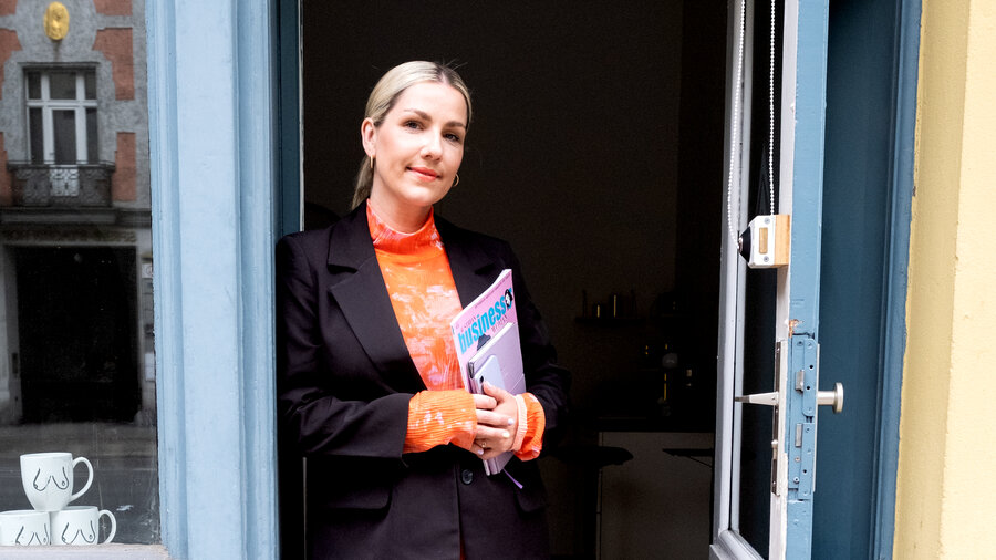 Eine blonde Frau mit orangfarbenem Kleid und dunkler Jacke lehnt in einer Tür.