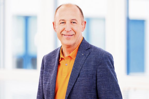 Mann mit grauem Sakko und orangefarbenem Poloshirt vor unscharfem Hintergrund mit Fenstern