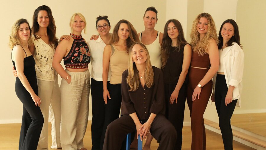 Ein Team an Frauen steht in einem Kursraum. Vor der Gruppe sitzt eine Frau auf einem Hocker. Alle lachen entspannt in die Kamera