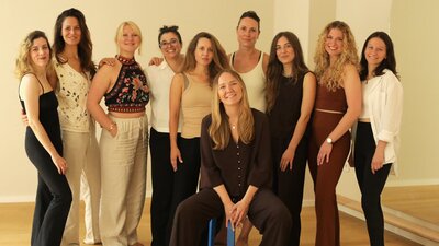 Ein Team an Frauen steht in einem Kursraum. Vor der Gruppe sitzt eine Frau auf einem Hocker. Alle lachen entspannt in die Kamera