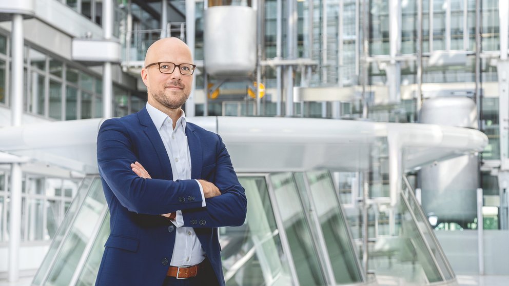 Auf dem Bild ust ein Mann mit Brille und blauem Anzug zu sehen, der in einer futuristisch anmutenden Halle steht.