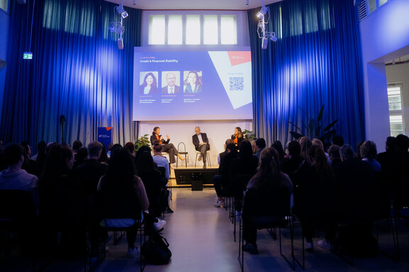 Drei Personen sitzen auf einer Bühne vor einem Publikum in einem Raum mit blauen Vorhängen und einer großen Projektionsfläche mit Text und Bildern.