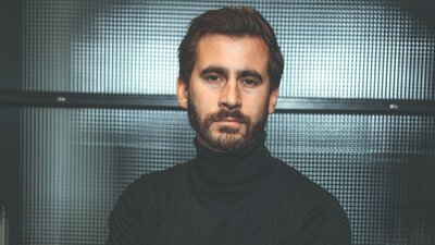 Zu sehen ist ein junger Mann in dunklem Pullover. Er verschränkt seine Arme vor der Brust, hat kurze dunkle Haare und einen dunklen Bart und blickt neutral in die Kamera.