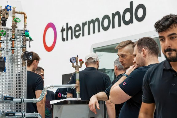 Mehrere Personen in schwarzer Arbeitskleidung vor einer Wand mit dem Schriftzug 'thermondo' und technischen Rohrinstallationen.
