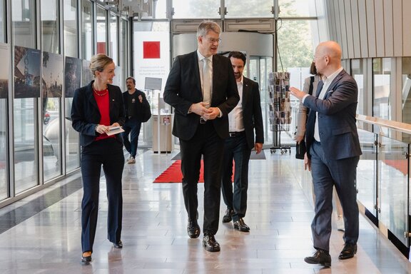 Zu sehen sind vier seriös gekleidete Personen, die in einer Art Lobby nebeneinander herlaufen und sich dabei unterhalten.