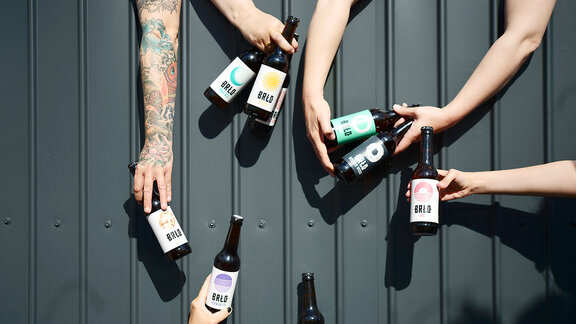 Sechs Hände halten verschiedene BRLO Bierflaschen mit bunten Etiketten vor einer dunklen Metallwand
