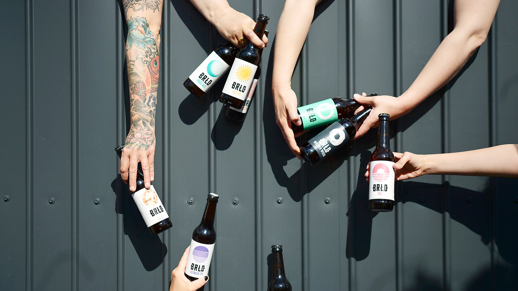 Sechs Hände halten verschiedene BRLO Bierflaschen mit bunten Etiketten vor einer dunklen Metallwand