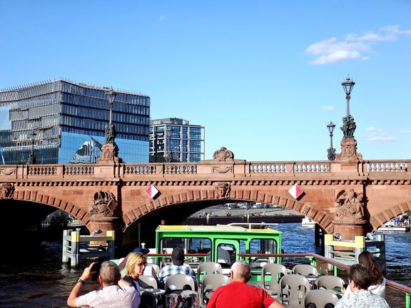 Blick auf eine Brücke mit zwei Statuen und Fahnen, im Vordergrund ein Boot mit grüner Kabine, im Hintergrund moderne Gebäude.