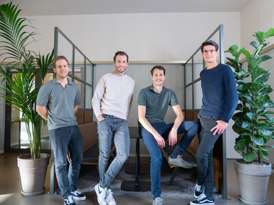 Vier junge Männer stehen nebeneinander in einem modern eingerichtetem Büro. Alle tragen blaue und grüne Töne. Sie lächeln in die Kamera