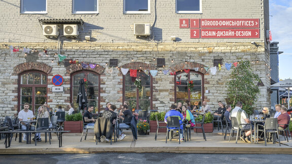 Ein Backsteingebäudemit bunter Beleuchtung und Dekoration. Davor sitzen Menschen in Sitzgruppen.  