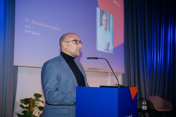 Mann im Anzug steht an einem blauen Rednerpult vor einer Leinwand mit Text 'Dr. Sebastian Schäfer CEO HoFT Berlin'.