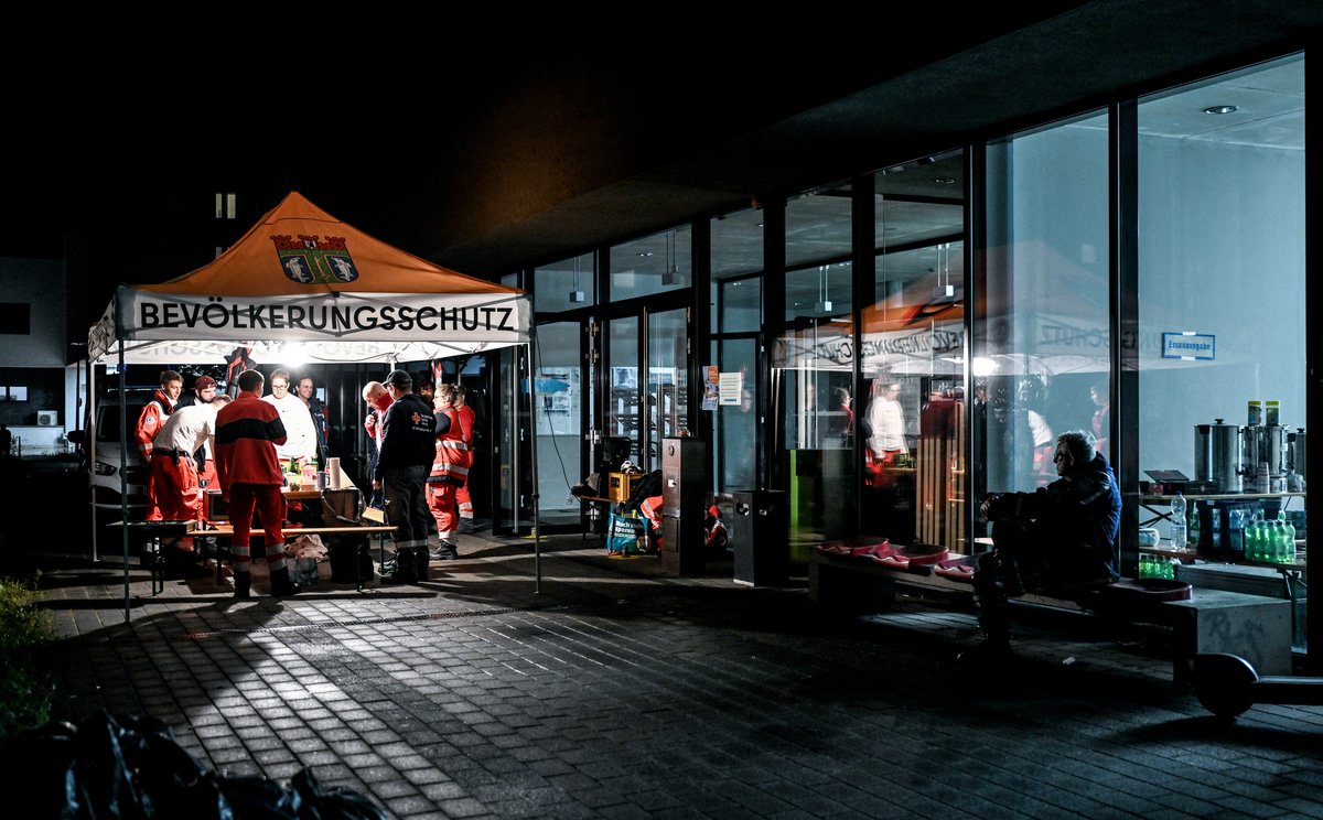 Gruppe von Einsatzkräften unter einem Zelt mit der Aufschrift 'BEVÖLKERUNGSSCHUTZ' bei Nacht vor einem Glasgebäude