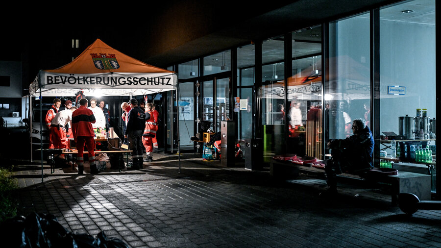 Gruppe von Einsatzkräften unter einem Zelt mit der Aufschrift 'BEVÖLKERUNGSSCHUTZ' bei Nacht vor einem Glasgebäude