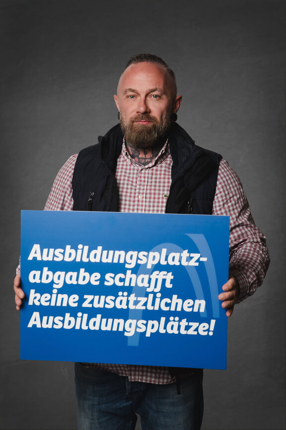 Eine Person steht vor einem neutralen grauen Hintergrund und hält ein großes blaues Schild mit der weißen Aufschrift: "Ja zur Ausbildung. Nein zur Abgabe!"