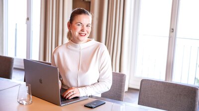 Eine Frau sitzt an einem hellen Konferenztisch und lächelt in die Kamera. Vor ihr steht ein Computer und sie trägt einen hellen Pullover