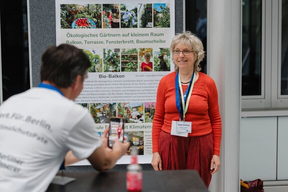 Zu sehen ist eine rotgekleidete Frau an einem Stand, die sich mit einem Interessierten unterhält.