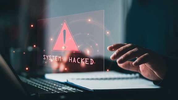 Zu sehen ist ein Laptop und eine Hand über der Tastatur. Hologramartig schwebt die Benachrichtigung "System Hacked" - also System gehackt über der Tastatur. 