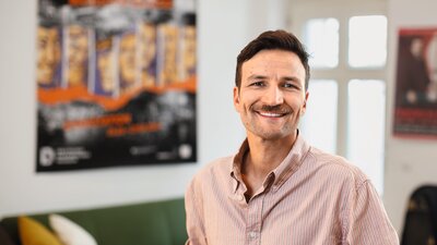 Zu sehen ist ein junger Mann, mit kurzen dunklen Haaren und einem hellen Hemd. Er schaut lächelnd in die Kamera.