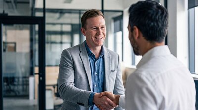 Zu sehen ist ein Mann im Anzug, der die Hand eines Geschäftspartners oder Kollegen schüttelt, der von hinten zu sehen ist.