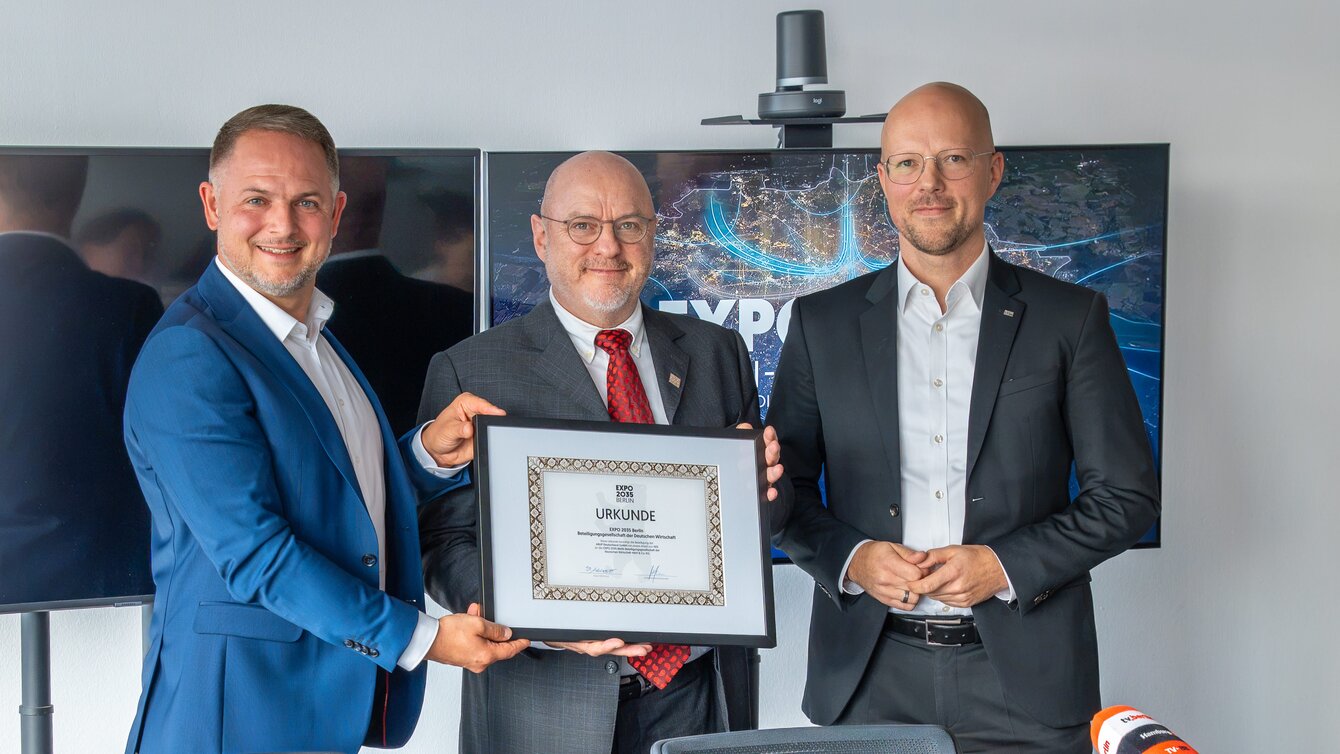 Auf dem Bild sind drei Männer zu sehen. Sie stehen vor zwei Bildschirmen und halten ein gerahmtes Zertifikat mit der Aufschrift "EXPO 2035" in die Kamera.