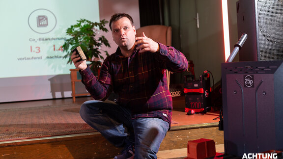 Ein Mann in einem karierten Hemd sitzt vor einer Leinwand auf einer Treppenstufe und gestikuliert. Es ist Roman Avanius von 25p