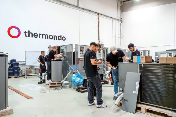 Mehrere Personen arbeiten in einer Werkhalle mit technischen Geräten und Bauteilen, an der Wand das Logo von Thermondo.