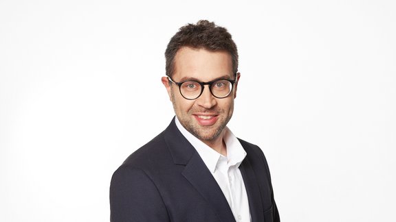 Mann mit kurzen braunen Haaren trägt weißen Hemdkragen und schwarzen Anzug, steht vor hellem Hintergrund.