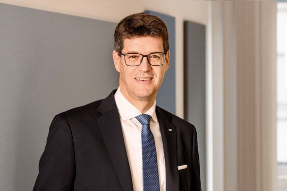 Mann in einem schwarzen Anzug mit weißem Hemd und blauer Krawatte, steht vor einer neutralen Wand mit grauen Paneelen.